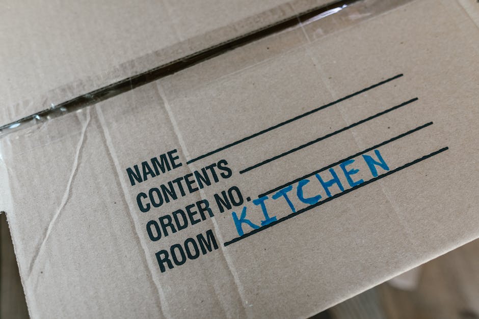 Close-up of a brown cardboard moving box with printed labels indicating sections for 'Name', 'Contents', 'Order No.', and 'Room', with 'Kitchen' handwritten in blue marker next to the 'Room' label. Th