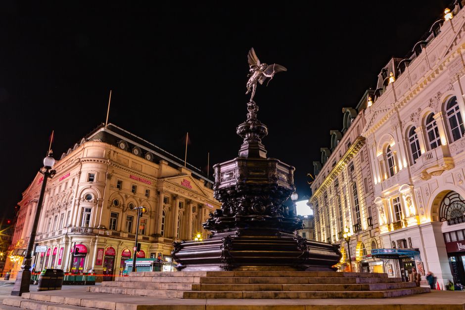 Nighttime scene at Piccadilly Circus showing the Shaftesbury Memorial Fountain with the illuminated statue of Anteros on top, surrounded by historic buildings with ornate facades and large windows. Th