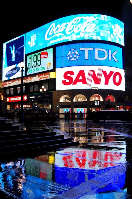 Nighttime scene at Piccadilly Circus showing the Shaftesbury Memorial Fountain with the illuminated statue of Anteros on top, surrounded by historic buildings with ornate facades and large windows. Th