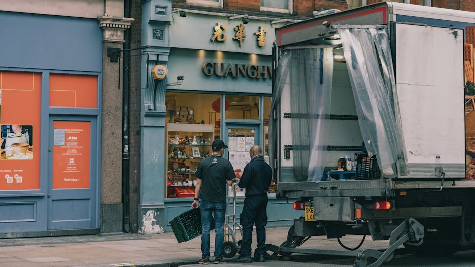 View of a narrow residential street in Soho W1 with red brick buildings and white sash windows, featuring street signs for Balderton Street and Providence Court. In the alleyway, two movers wearing da